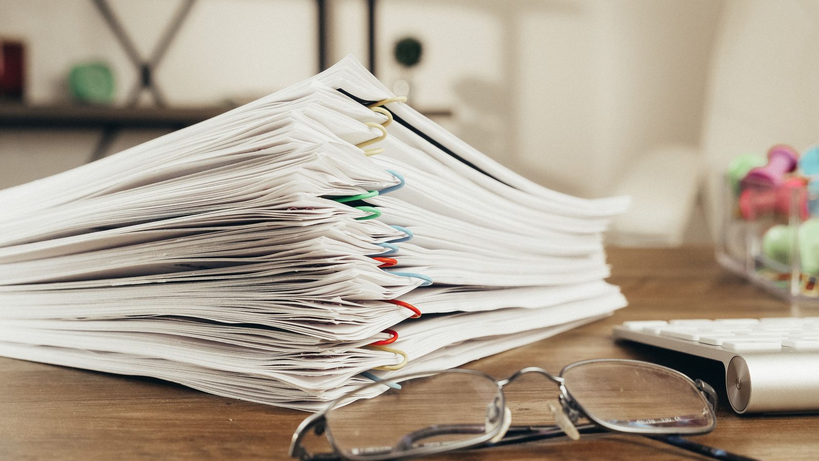 A tall stack of paper-clipped documents beside reading glasses on a desk.